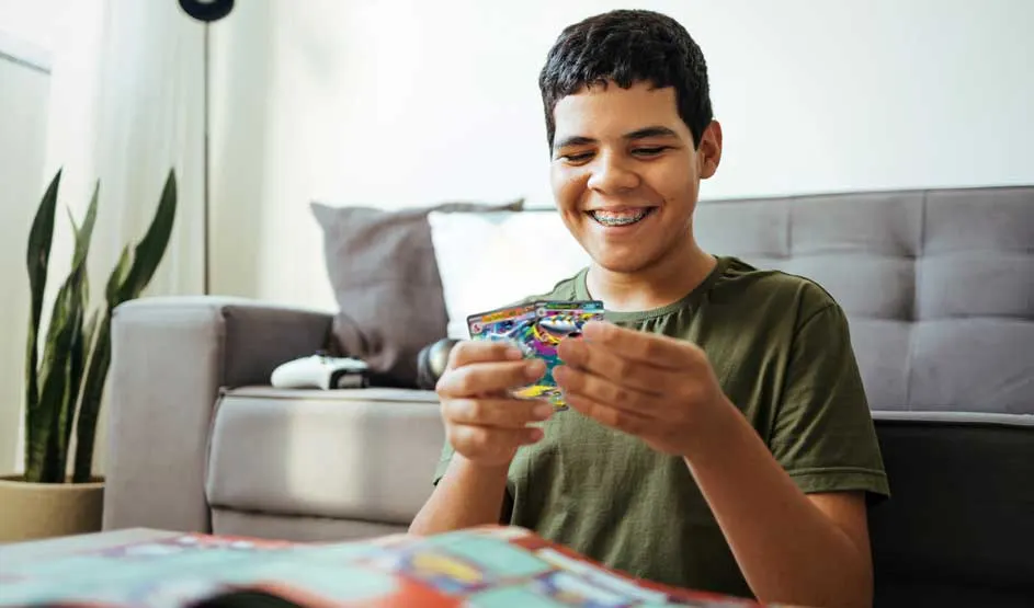 Un garçon souriant est assis dans le salon et regarde avec enthousiasme des cartes colorées, avec un canapé et une plante derrière.