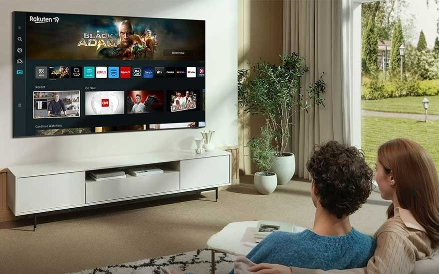 Een woonkamer met een tv die "Black Adam" toont, een witte tv-meubel, planten en een groot raam.