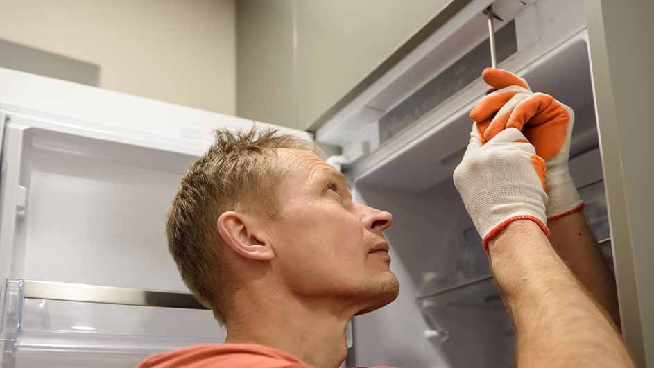 A person in gloves uses a screwdriver to fix the white refrigerator. The background shows a light-colored wall.