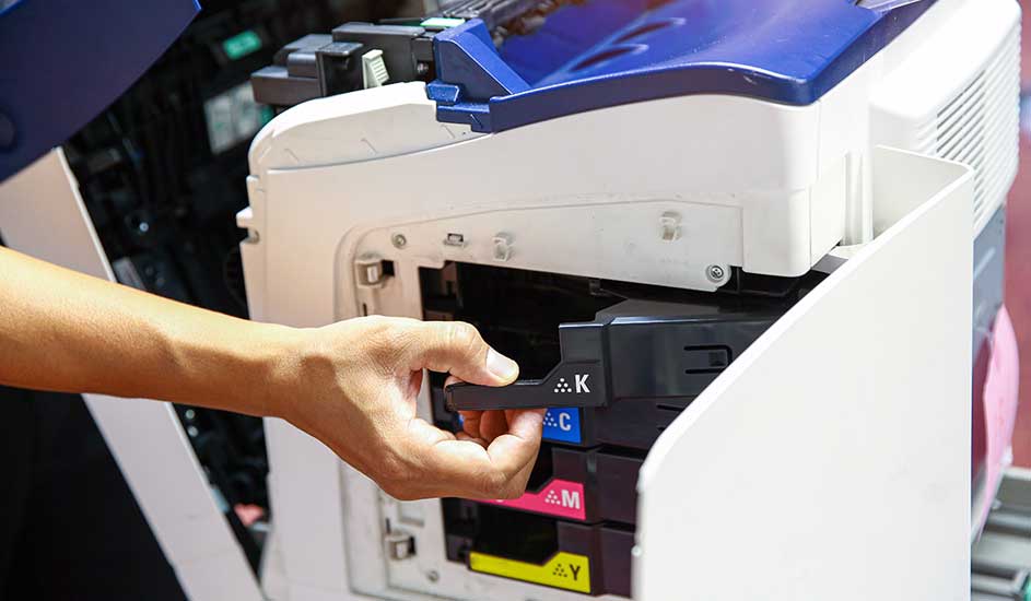 Een hand vervangt een zwarte cartridge in een witte printer met blauwe accenten. Kleurcartridges zitten eronder.