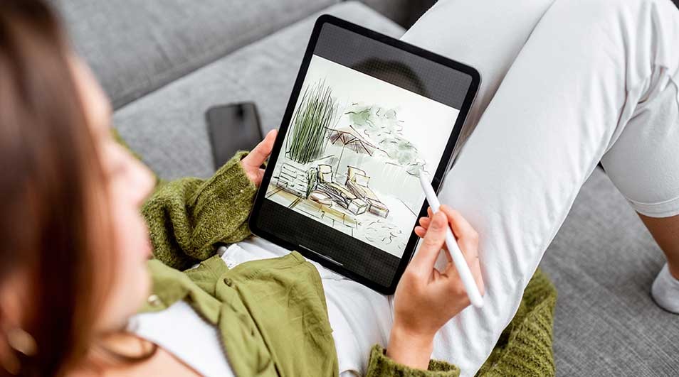 Vue de dessus d'une tablette affichant un dessin de meubles d'extérieur, tenue par une main avec un stylet sur un canapé gris.