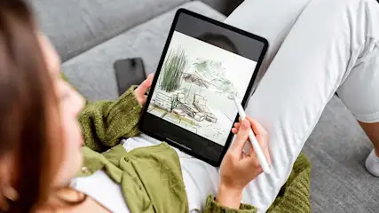 Vue de dessus d'une tablette affichant un dessin de meubles d'extérieur, tenue par une main avec un stylet sur un canapé gris.