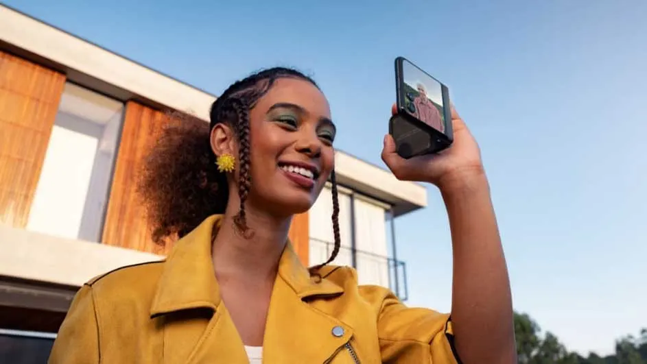 Une personne en veste jaune prend un selfie avec un téléphone devant un bâtiment moderne et un ciel bleu.