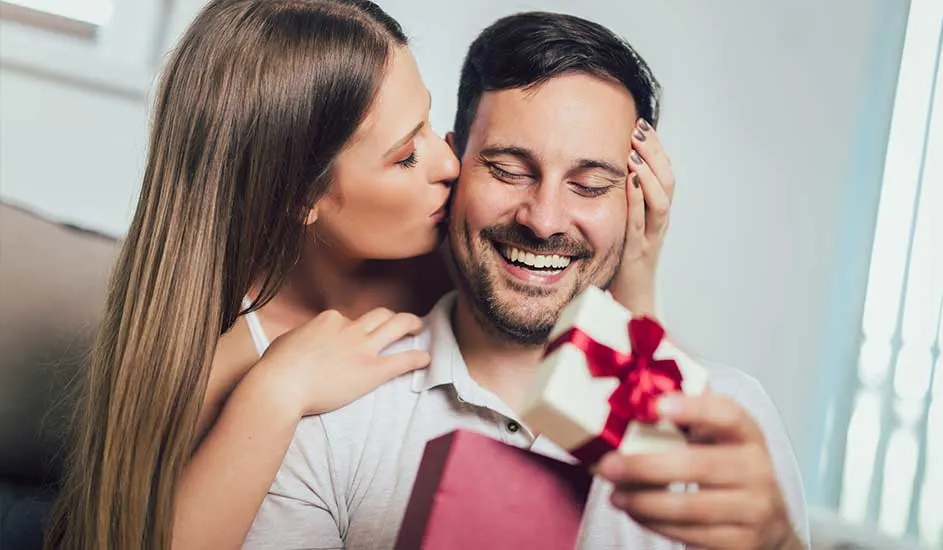 Un homme reçoit un cadeau avec un ruban rouge, une femme l'embrasse. L'arrière-plan est blanc avec une fenêtre.