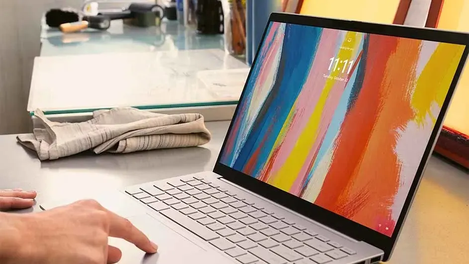 Een laptop staat open op een bureau met een kleurrijk scherm. Er ligt een doek en kantoorbenodigdheden op de achtergrond.