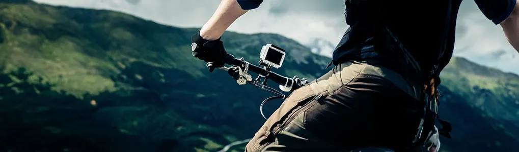 Bike with camera, black gloves, and dark pants on a mountain path. Background shows mountains and cloudy sky.