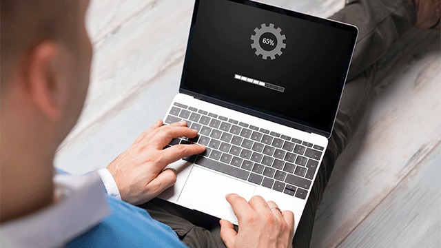 Laptop toont 65% update met tandwiel. Persoon typt op toetsenbord. Grijze achtergrond.