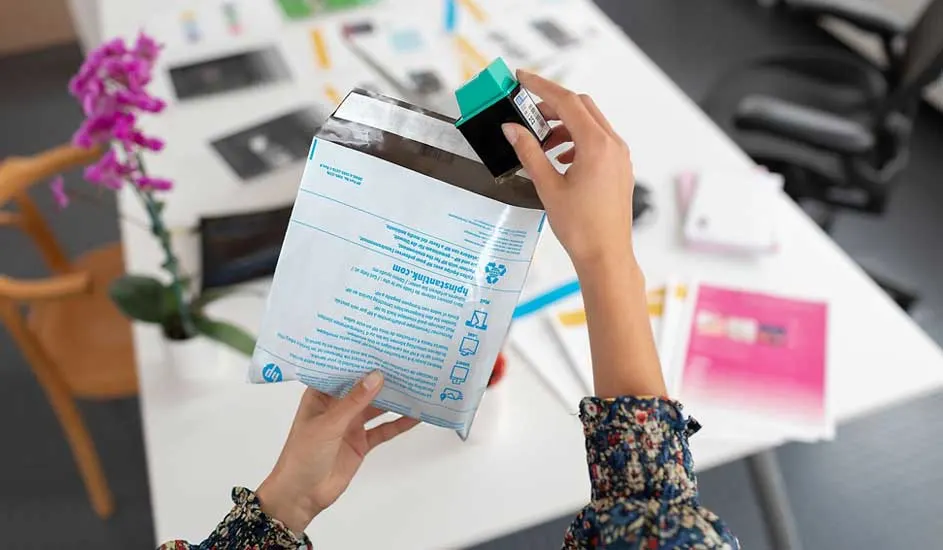 Een persoon houdt een HP-envelop en een cyaan printercartridge vast boven een witte tafel. Er staan bloemen op de tafel.