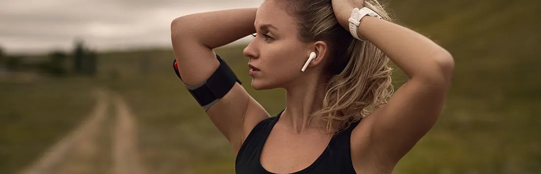 Femme avec brassard noir, écouteurs blancs et montre, mains derrière la tête. Sentier et végétation en arrière-plan.