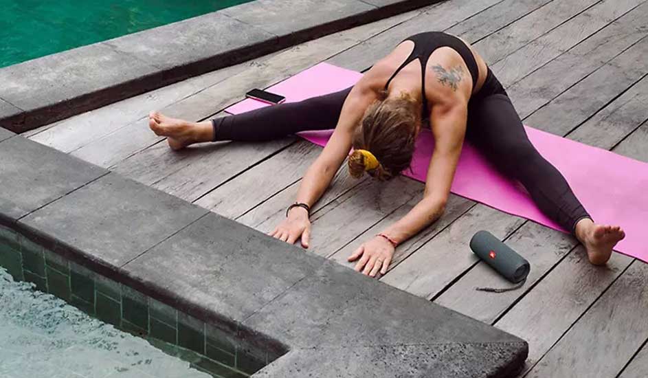 Une personne fait du yoga sur un tapis rose près d'une piscine. Un haut noir et un pantalon foncé sont portés. Un téléphone et un haut-parleur sont à proximité.