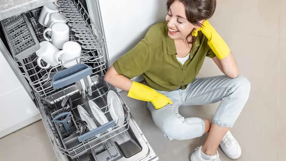 Un lave-vaisselle ouvert montre des tasses, des assiettes et des couverts blancs. Le sol est en carrelage clair.