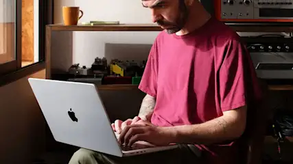 Un ordinateur portable argenté est utilisé devant une étagère en bois avec des équipements audio et une tasse orange.