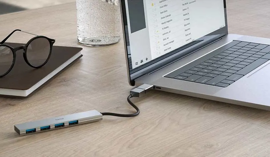 Een zilveren laptop is aangesloten op een USB-hub met vier blauwe poorten op een houten bureau. Een bril en notitieboek liggen ernaast.