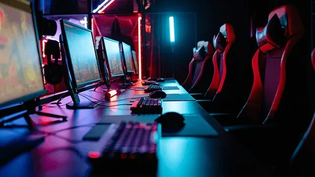 Een rij computers met schermen, toetsenborden en muizen op tafels, met rode gamingstoelen erachter, in een donkere kamer met neonverlichting.