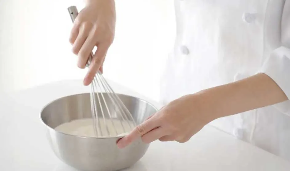Une personne en blouse blanche fouette un liquide clair dans un bol métallique sur une table blanche.