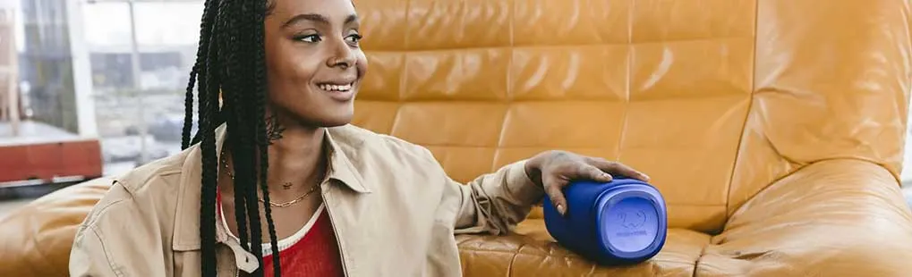 Une personne est assise sur un canapé jaune avec une enceinte Bluetooth JBL bleue à côté, portant une veste beige et un haut rouge.