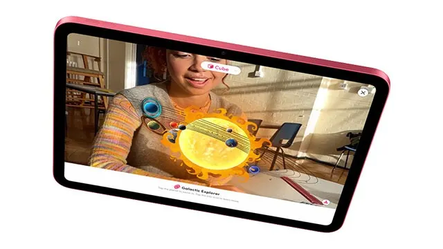 Een roze tablet toont een zonnestelsel-app in een lichte kamer. Stoelen en een bureau zijn zichtbaar op de achtergrond.