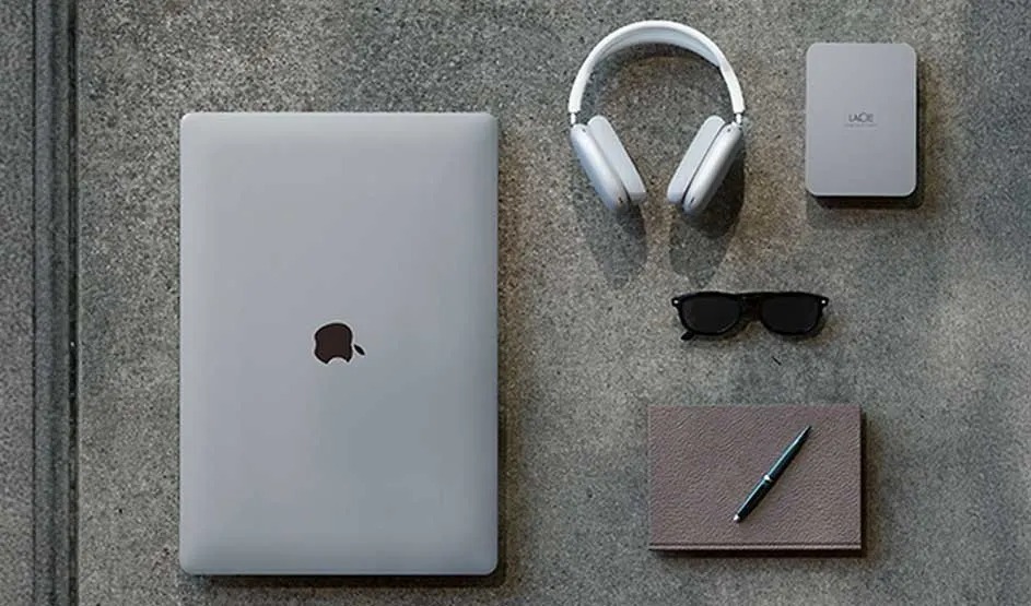 Een Apple MacBook, LaCie externe harde schijf, hoofdtelefoon, notitieboek en zonnebril liggen netjes op een tafel.