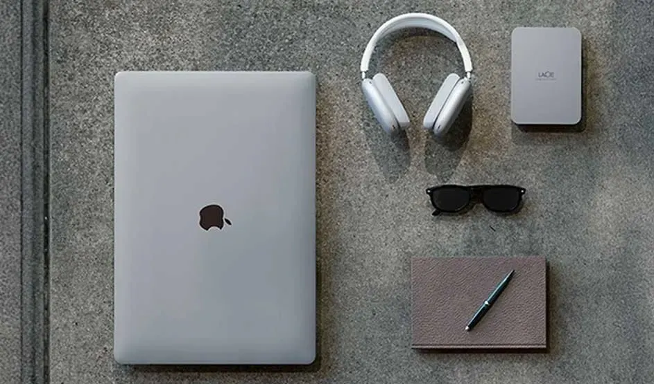 Een Apple MacBook, LaCie externe harde schijf, hoofdtelefoon, notitieboek en zonnebril liggen netjes op een tafel.