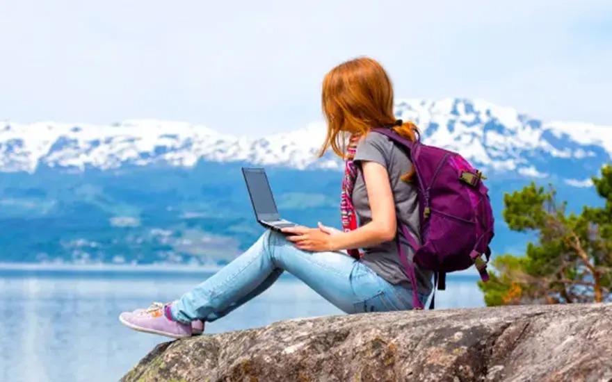 A laptop sits on a rock with a purple backpack. Mountains and a body of water are in the background.