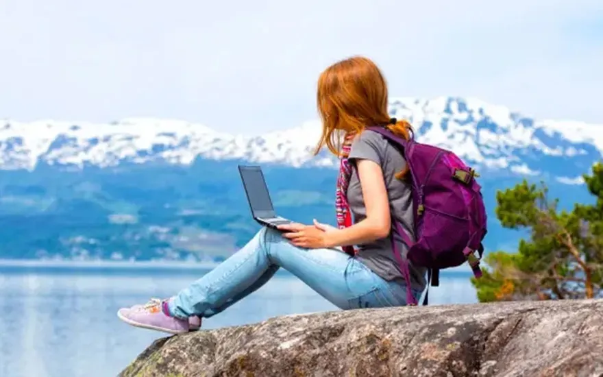 A laptop sits on a rock with a purple backpack. Mountains and a body of water are in the background.