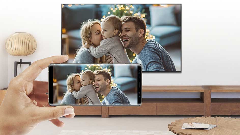 Hand houdt smartphone voor tv. Beide schermen tonen foto van familie. Houten kast en lamp in de kamer.