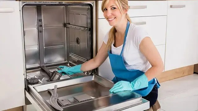 A person wearing blue gloves and an apron is cleaning the inside of an open dishwasher. No brand is visible.