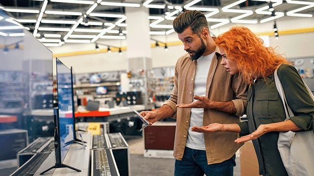 Dans un magasin, des téléviseurs noirs sont exposés sur des étagères métalliques. L'éclairage du plafond est vif.