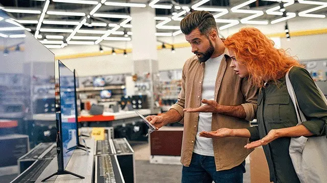 Dans un magasin, des téléviseurs noirs sont exposés sur des étagères métalliques. L'éclairage du plafond est vif.
