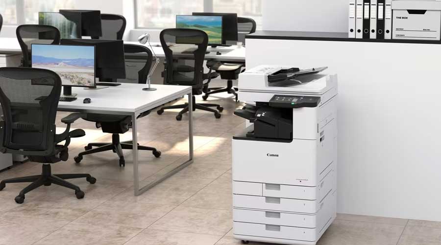 White Canon multifunction printer in a modern open-plan office with desks, chairs and monitors in the background.