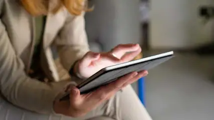 Une personne tient une tablette et touche l’écran, assise dans un intérieur calme aux couleurs neutres et à l’arrière-plan flou.