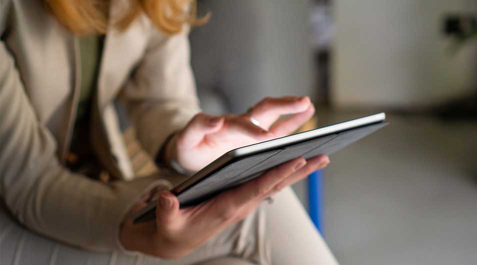 Een persoon houdt een tablet vast en raakt het scherm aan, zittend in een rustige binnenruimte met zachte focus en neutrale kleuren.