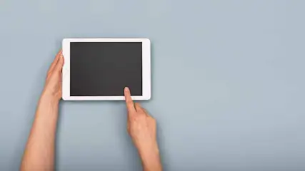 Une tablette blanche à écran noir est tenue et touchée par une main sur un fond bleu clair.