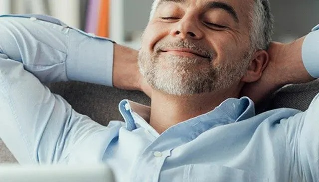 Un homme en chemise bleue se détend, les mains derrière la tête, sur un canapé gris, avec des livres en arrière-plan.