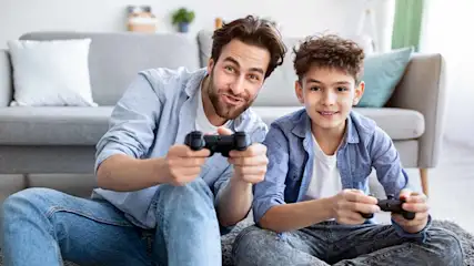 Een grijze sofa met kussens op een tapijt. Twee mensen spelen computerspelletjes met zwarte controllers.