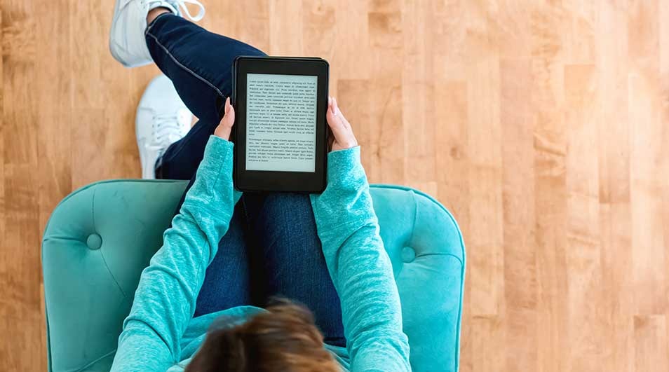 A person in jeans and white shoes sits in a turquoise chair, reading a black e-reader against a light wood floor.