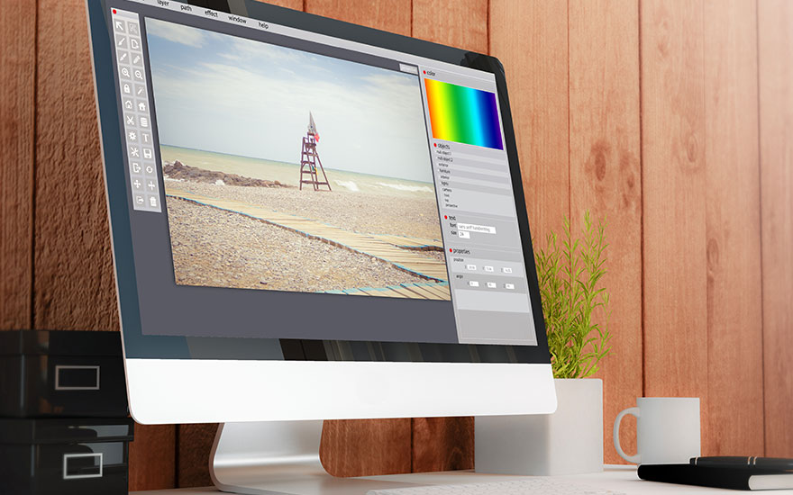 Écran affichant un logiciel d'édition avec une image de plage, palette de couleurs, boîtes noires, plante, tasse blanche et stylo sur un bureau.