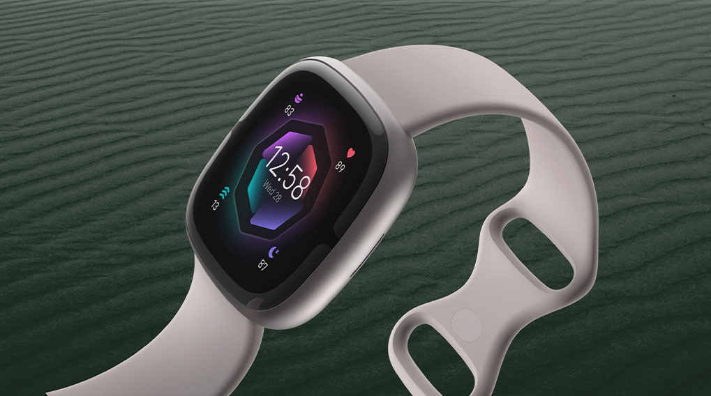 Een grijze smartwatch toont 12:58 op een kleurrijk scherm, liggend op een groene achtergrond met textuur.