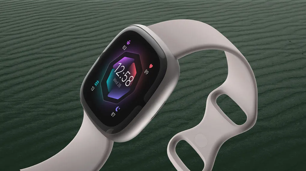 Een grijze smartwatch toont 12:58 op een kleurrijk scherm, liggend op een groene achtergrond met textuur.