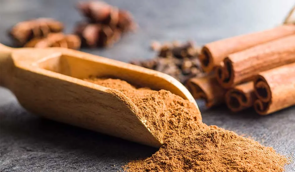 Une cuillère en bois remplie de poudre de cannelle, avec des bâtons de cannelle, de l'anis étoilé et des clous de girofle en arrière-plan.