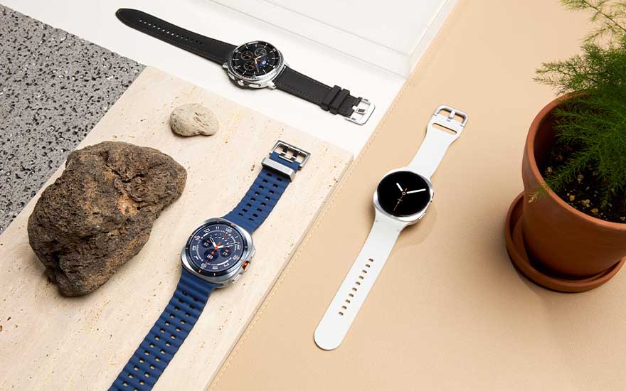 Drie Samsung-smartwatches met zwarte, blauwe en witte bandjes liggen op een stijlvolle tafel met stenen en een plant in pot.