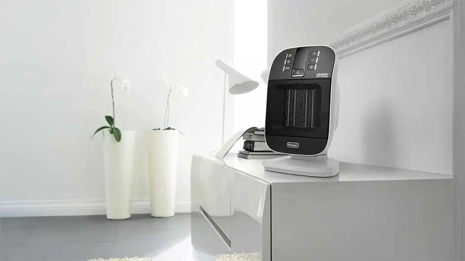 A black and white heater sits on a white cabinet. Two white vases with flowers stand nearby in a white room with a white lamp.