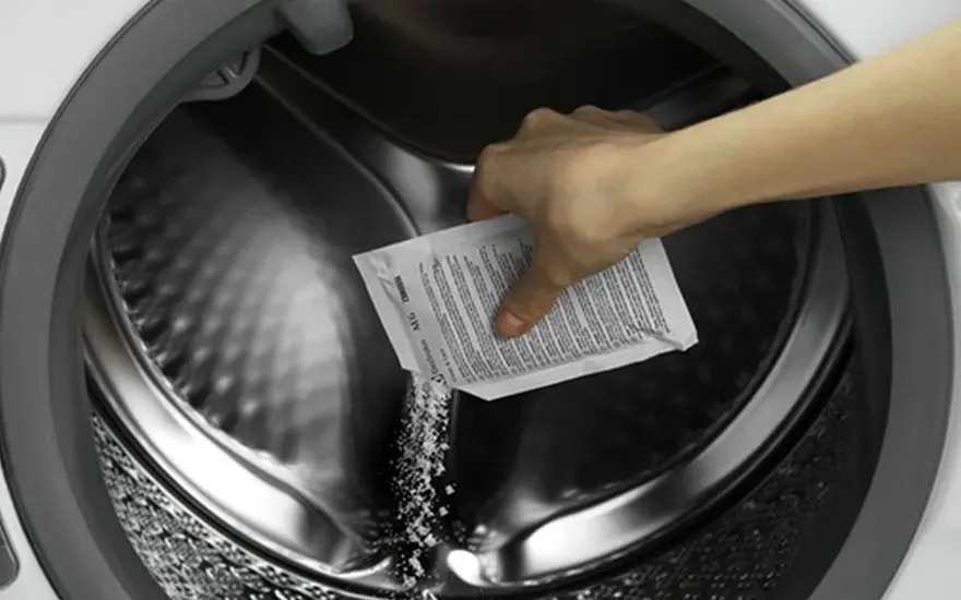 Une main verse de la poudre d’un sachet dans le tambour d’un lave-linge ouvert, la marque du lave-linge n’est pas visible.