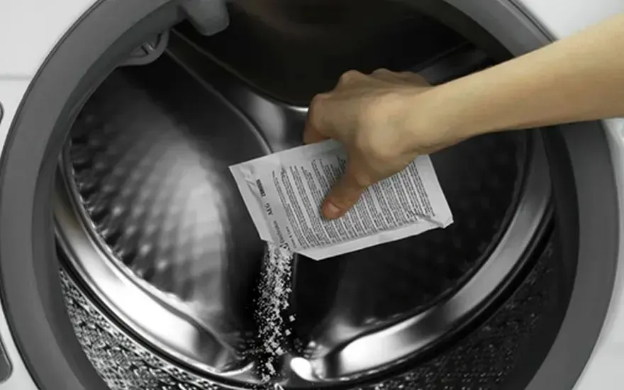 Een hand giet poeder uit een zakje in de trommel van een open wasmachine, merk van de wasmachine is niet zichtbaar.