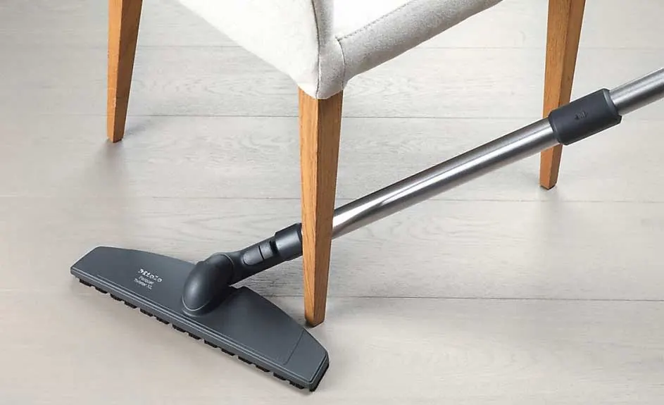 Un aspirateur gris nettoie sous une chaise beige aux pieds en bois clair sur un plancher en bois blanchi.