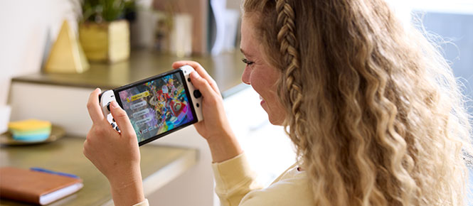 Een Nintendo Switch-console met een kleurrijk spel wordt in een kamer gehouden. Op een tafel staan decoratieve objecten.