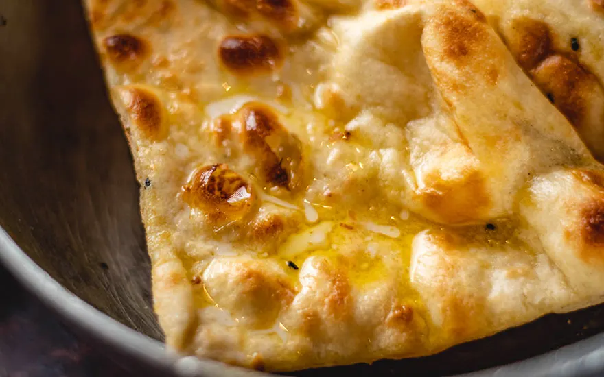 Gros plan d'un pain naan doré et beurré sur une assiette grise. Le pain est brillant et cuit de manière inégale.