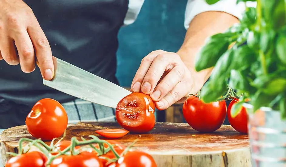 Een kok snijdt tomaten op een houten plank met een mes. Naast de tomaten staat een basilicumplant.