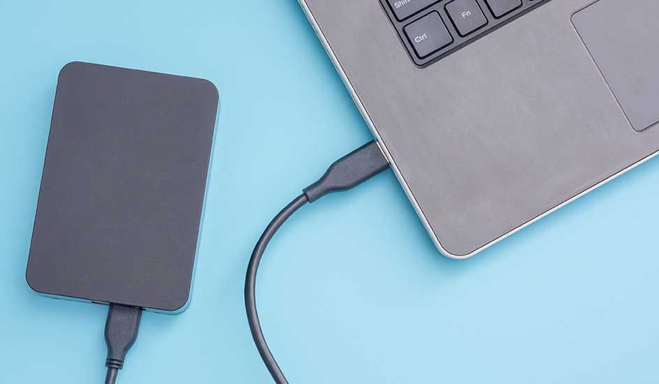Un disque dur externe gris est connecté à un ordinateur portable gris avec un câble sur un fond bleu clair.
