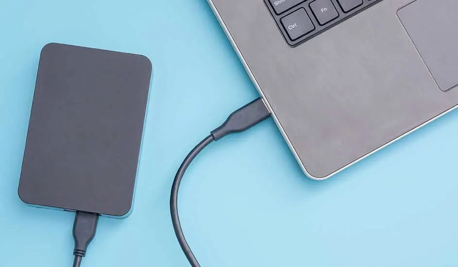 Un disque dur externe gris est connecté à un ordinateur portable gris avec un câble sur un fond bleu clair.